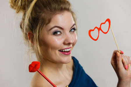 Happy blonde woman holding carnival accessories on stick fake red lips and paper heart shaped glasses, having fun. On grey wall backgroundの写真素材