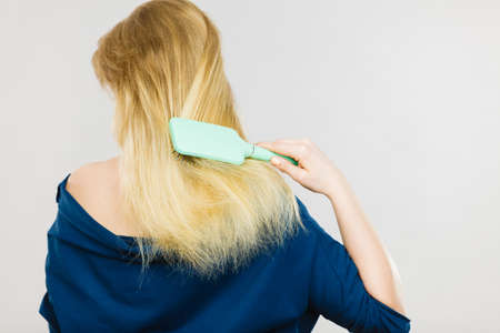 Everyday hygiene and care about good look. Back view of blonde casual girl combing her long straight hair. Woman using blue comb hairbrush.の写真素材
