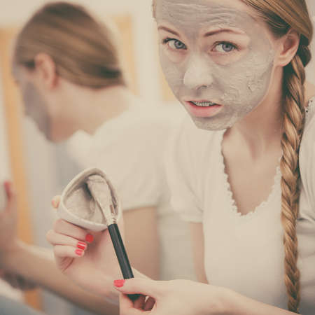 Skincare. Blonde woman in bathroom with gray clay mud mask on her face. Young lady taking care of skin. Spa beauty wellness. Toned imageの写真素材