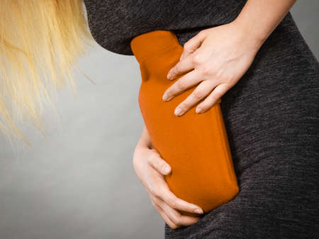 Unrecognizable woman having strong stomach ache. Female suffer on belly pain, holding hot red water...