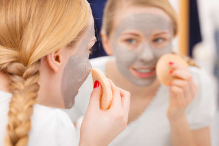 Beauty procedures spa and skin care concept. Young woman looking in mirror, removing facial clay mud mask with sponge in bathroomの写真素材