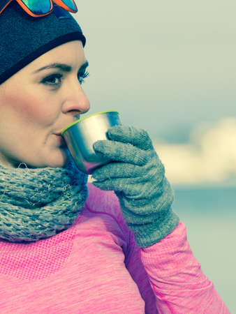 Woman athlete fit girl wearing warm sporty clothes outdoor in cold weather drinking hot tea from vacuum flask thermos, warming up. Sports, activities, hiking in winter or autumn timeの写真素材
