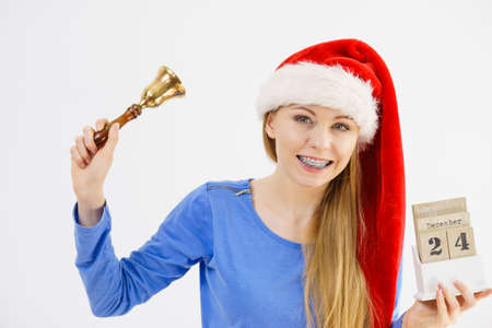 Happy positive young teenage woman wearing Santa Claus christmas hat holding calendar with 24 december date and bellの写真素材
