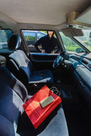 Anti theft system problem concept. Burglar thief man wearing black clothes breaking into car, stealing smartphone and red shopping bagの写真素材
