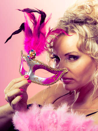 Holidays, people and celebration concept. Woman blonde mid aged model holding in hand pink carnival mask, feather fan on bright backgroundの写真素材
