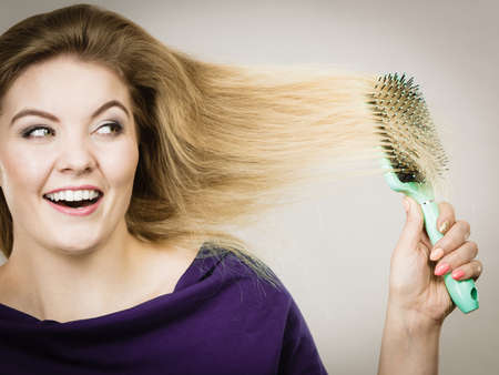Happy woman combing her hair with brush. Young smiling female with natural blond straight long hairs, studio shot on greyの写真素材