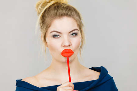 Funny woman holding big red lips on stick. Blonde young female ready for party, on grey background. Humor costume, smart and carnival concept.の写真素材
