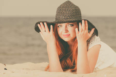 Summertime outfit accessories concept. Happy redhead woman wearing big black sun hat lying on beach during summer.の写真素材