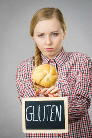 Young woman with braided hair holding small black board with gluten sign and bun bread roll. Bakery and bread allergy problem.の写真素材