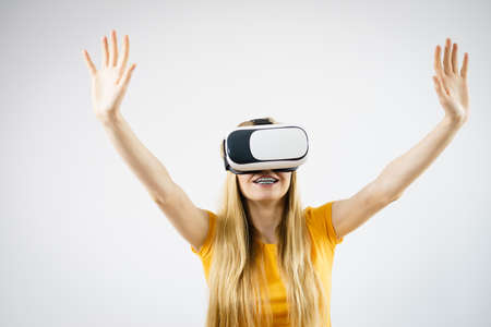 Young woman wearing virtual reality goggles headset, vr box, stretching arms. Studio shot on gray....