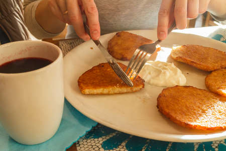 Delicious potato pancakes on plate with sour cream and tea in mug. Traditional slavic dish. Woman hands holding cutleryの写真素材