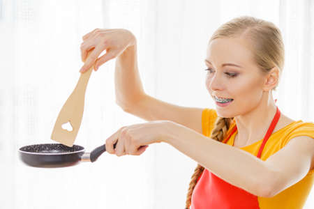 Happy cheerful young woman wearing apron holding small cooking pan and wooden spatula about to cook in kitchen.の写真素材