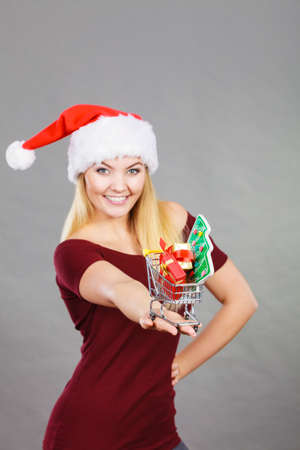 Xmas, seasonal sales, winter celebration concept. Happy woman wearing Santa Claus helper hat holding shopping basket cart with little christmas tree and gifts inside.の写真素材