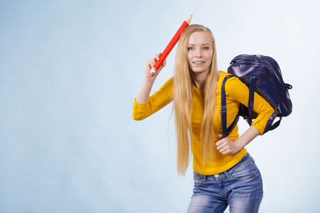Happy young blonde teenage girl going to school or college wearing backpack and holding big pencil.の写真素材
