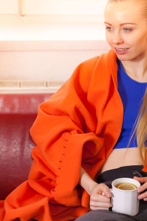 Young woman lying on sofa under warm blanket drinking tea, coffee or different hot drink from mug enjoying her leisure time during cold weather.の写真素材