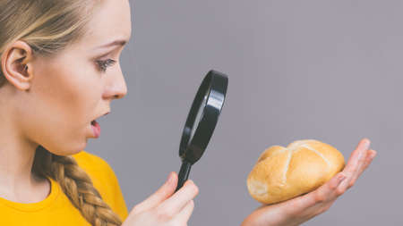 Shocked woman holding bun bread roll and magnifying glass examine wheat product ingredients seeing something disgustingの写真素材