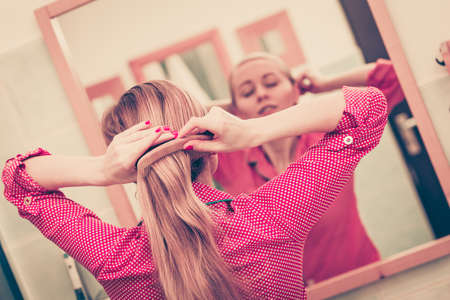 Woman combing brushing her long blonde smooth hair in bathroom, looking in mirror. Teen girl taking care refreshing her hairstyle in morning. Haircare concept.の写真素材