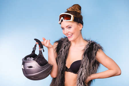 Winter sport activity concept. Atractive smiling girl wearing black bra, ski goggles and furry waistcoat holding helmet, blue background studio shot.の写真素材