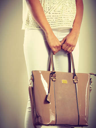 Fashion concept. Clothing and accessories. Woman hands holding beige handbag. Lady in white clothing with lacquered leather bag.の写真素材