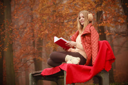 Autumn time education concept. Blonde girl with book. Attractive woman sitting on blanket in the park relaxing, foggy misty dayの写真素材