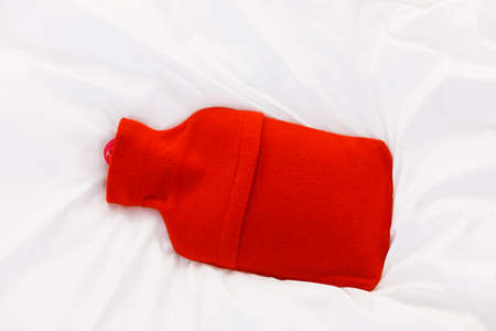 Useful things during fever concept. Warm red hot water bottle on white bedding