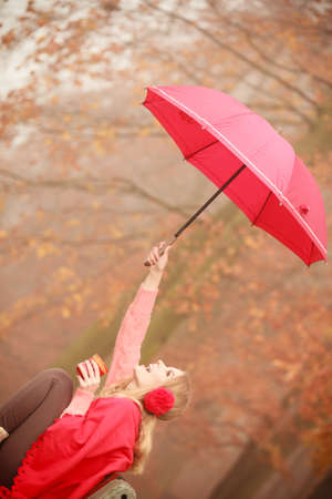 Happiness carefree and fall concept. Joyful woman relaxing in autumn park on bench under umbrella enjoying hot drink holding mug with warm beverage. Orange leaves backgroundの写真素材