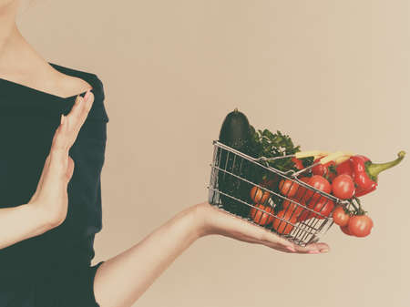 Adult woman do not like to eat vegetables, healthy food, vegetarian products. Female holding small shopping basket with green red vegetables, stop gesture, on greyの写真素材