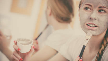 Skincare. Blonde woman in bathroom with gray clay mud mask on her face. Young lady taking care of skin. Spa beauty wellness. Filtered photoの写真素材