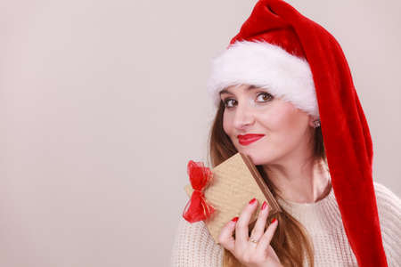 Cheerful woman wearing santa claus hat holding golden gift box with red bow. Christmas time, giving and happiness concept.の写真素材