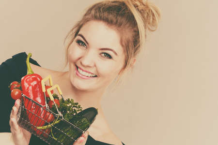 Buying good food, vegetarian products. Positive woman holding shopping basket with green red vegetables inside, recommending healthy high fibre diet, lifestyle modification, on greyの写真素材
