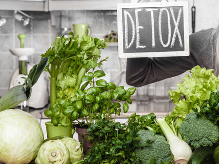 Woman in kitchen having many green vegetables presenting board with detox sign.の写真素材