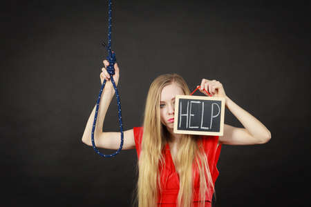 Suicidal skinny woman wearing elegant red dress holding help sign on dark board next to hanging rope with knot.の写真素材
