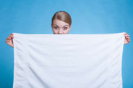 Workout, hygiene concept. Smiling woman holding a big white clean towel and hiding behind it, studio shot on blue backgroundの写真素材