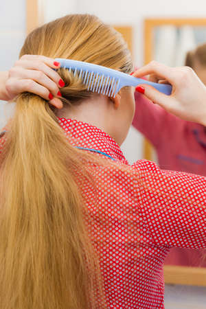 Woman combing brushing her long blonde smooth hair in bathroom, looking in mirror. Teen girl taking care refreshing her hairstyle in morning. Haircare concept.の写真素材