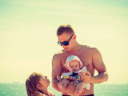 Toddler girl and father holding baby posing in summer clothes on sunny day with water in backgroundの写真素材