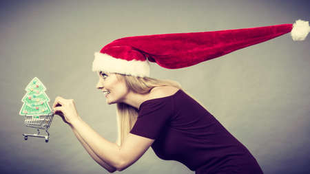 Xmas, seasonal sales, winter celebration concept. Happy woman wearing Santa Claus helper hat holding shopping basket cart with little christmas tree inside running for sale.の写真素材