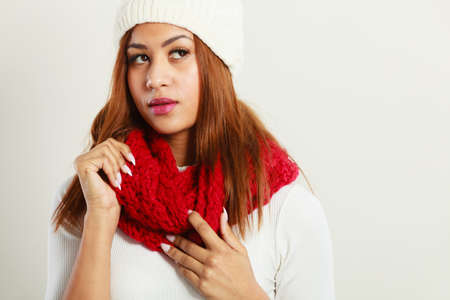 Winter clothing, fashion concept. Beautiful young mulatto woman wearing red woolen scarf white cap. Mixed race girl in wintertime clothes portrait, studio shotの写真素材