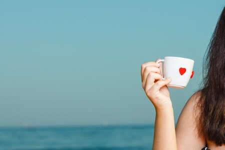 Woman hand holding white cup with red heart shape, blue sea and horizon in background.の写真素材
