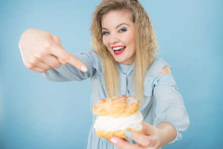 Sweet food and happiness concept. Funny joyful blonde woman pointing on yummy choux puff cake with whipped cream, excited face expression. On blueの写真素材
