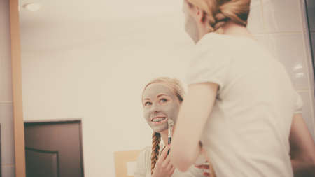 Skincare. Blonde woman in bathroom with gray clay mud mask on her face. Young lady taking care of skin. Spa beauty wellness. Filtered photoの写真素材