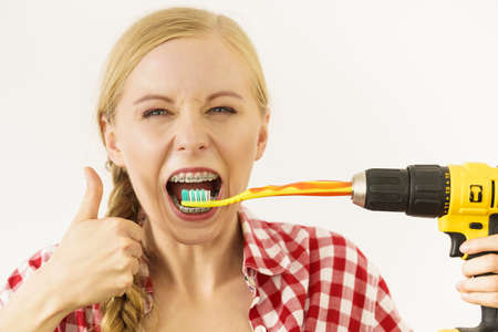 Funny teenage woman wearing braces holding drill about to brush her teeth.