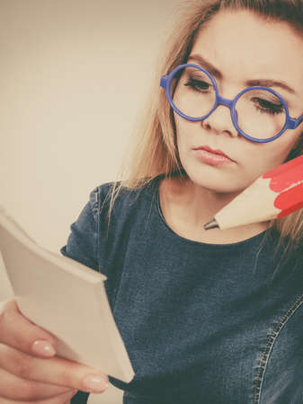 Serious student girl or female teacher, business coach holding huge red pencil and note paper, planning. Studio shot on greyの写真素材