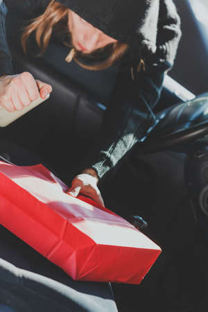 Anti theft system problem concept. Burglar thief man wearing black clothes breaking into car, stealing smartphone and red shopping bagの写真素材