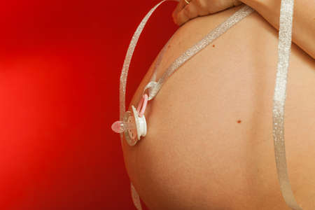 Parenthood, waiting for baby concept. Adult woman showing her big pregnant belly holding pacifier. Indoor shot on red backgroundの写真素材