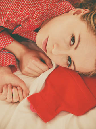 Woman lying on bed sleeping with warm red hot water bottle. Fever, flu treatment objects concept.の写真素材