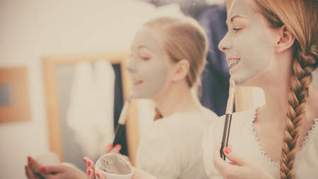 Skincare. Blonde woman in bathroom applying with brush gray clay mud mask to her face. Young lady taking care of skin. Spa beauty wellness.の写真素材