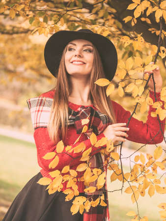 Autumn season colors in the park concept. Woman standing by the tree. Attractive lady wearing black hat and red sweather and very stylish checkered scarf.の写真素材