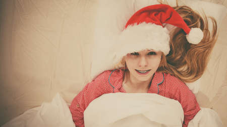 Happy woman waiting for Christmas season wearing pajamas and Santa Claus hat lying in bed.の写真素材