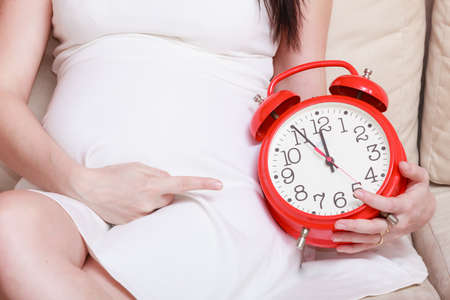 Pregnancy, motherhood, expecting concept. Pregnant woman holding big red old fashioned clock,...