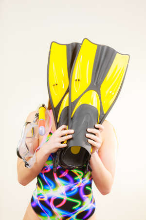 Woman wearing swimsuit covering her face with flippers, having fun studio shot on grey. Girl...